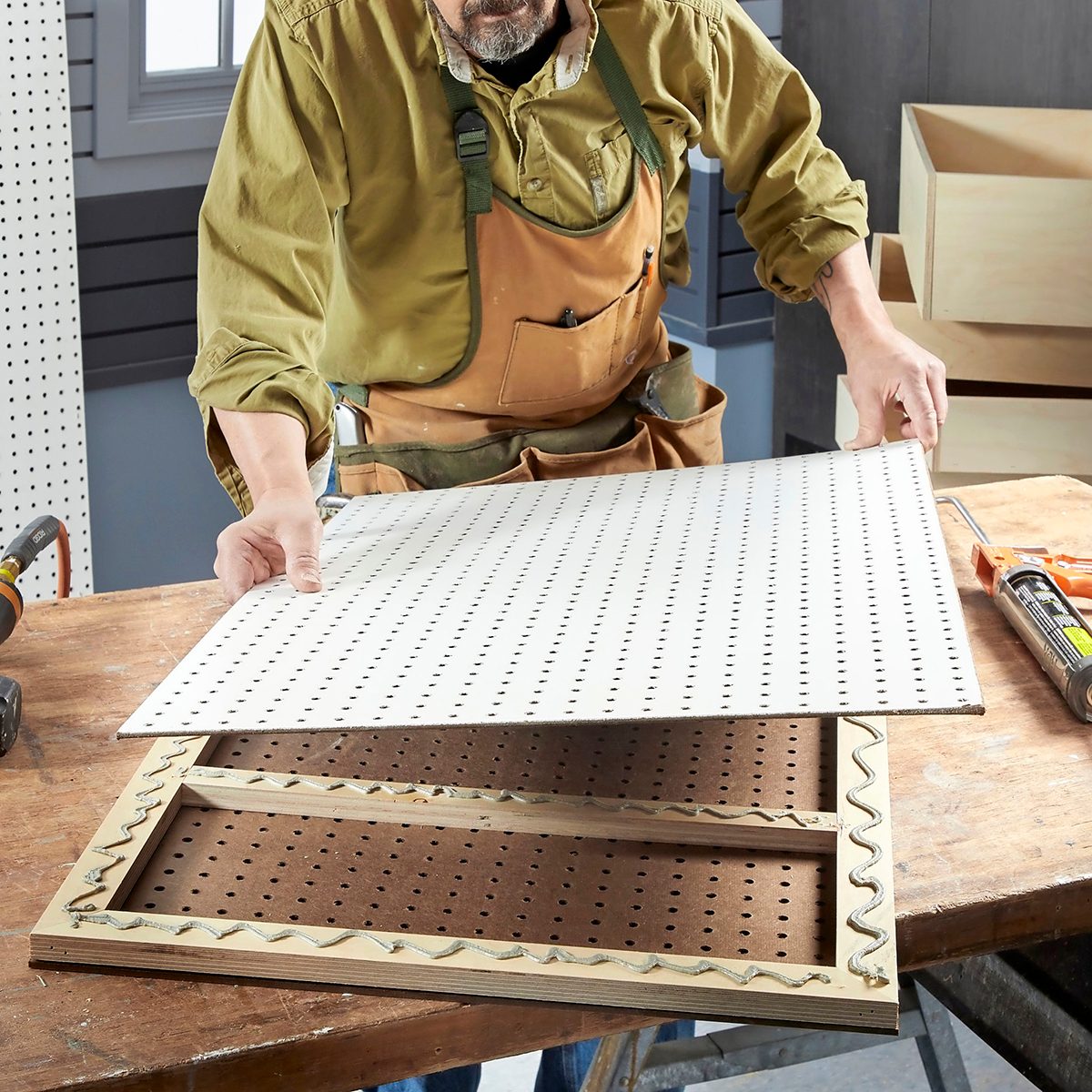 A person places a perforated board onto an assembled frame on a workbench, surrounded by tools and wooden boxes in a workshop setting.