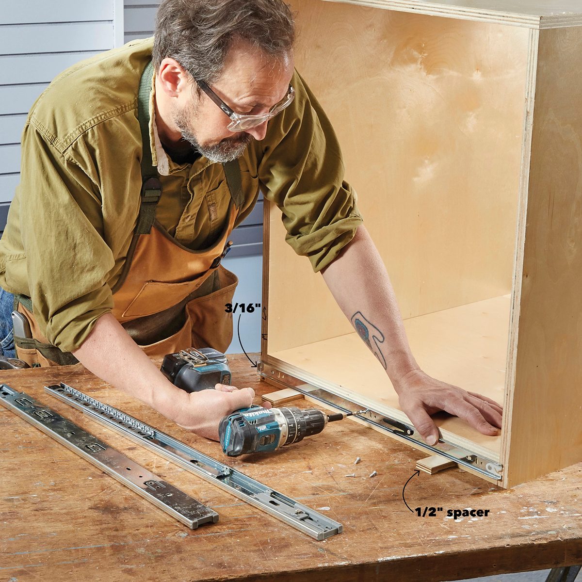 A man uses a power drill to attach a metal rail inside a wooden cabinet, with spacers and other tools nearby on a workbench.
