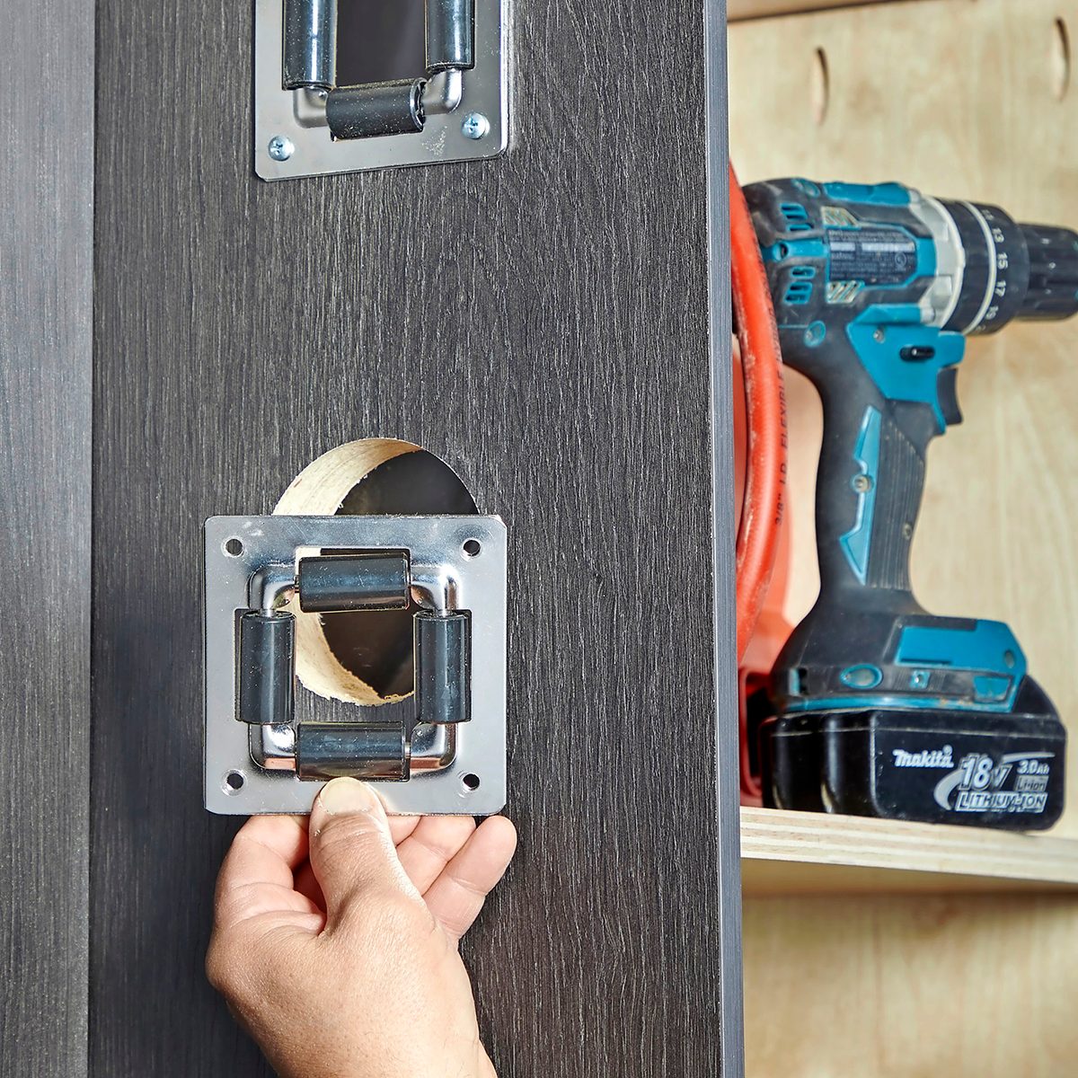 A hand positions a metal handle mechanism onto a wooden panel with a circular cutout, while a power drill rests nearby on a wooden shelf.