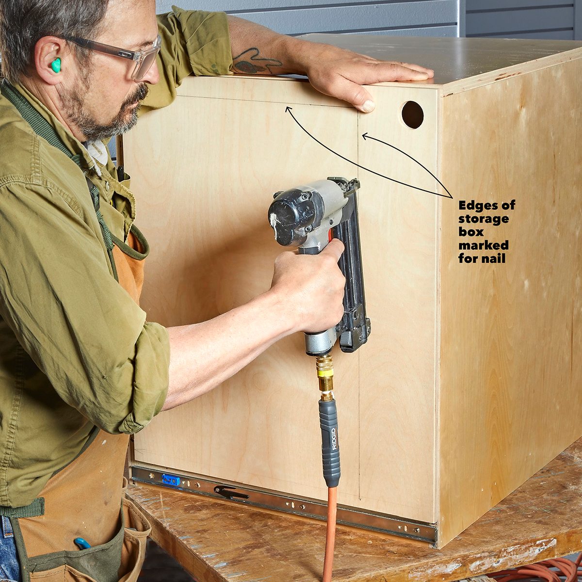 A person uses a nail gun to attach pieces of a wooden storage box, marked for nails, while working on a wooden table in a workshop.
