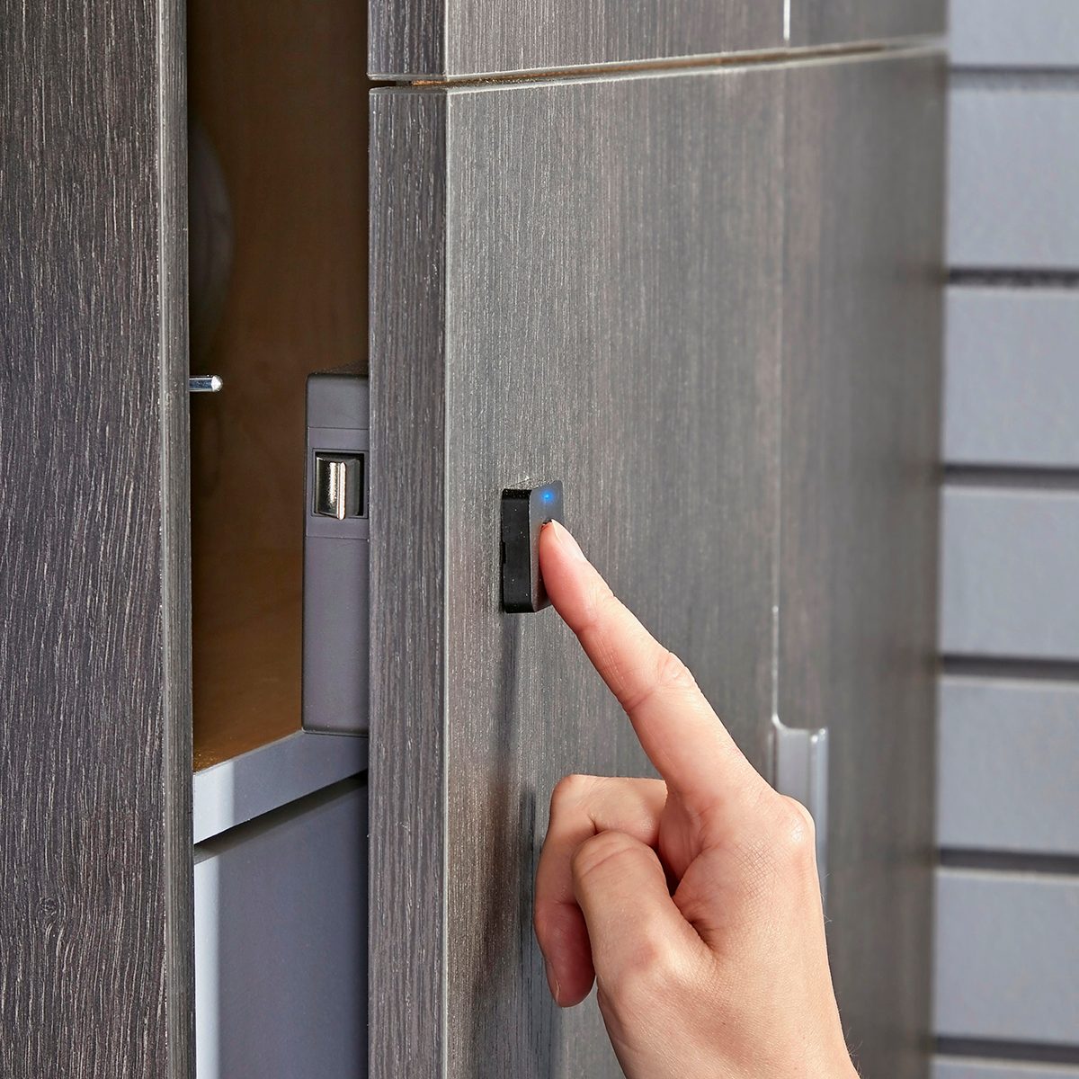 A hand presses a button on a gray cabinet door, which is partially open, revealing a USB hub and other items inside.