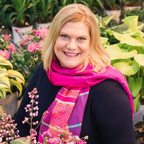 A woman with blonde hair and a pink scarf smiles among vibrant flowers and green foliage in a gardening setting.