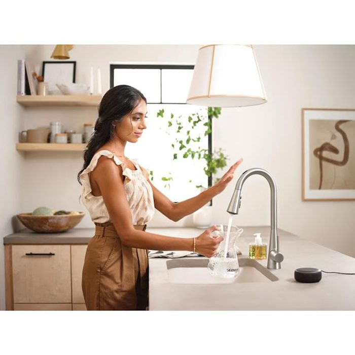 A woman pours water from a modern kitchen faucet into a glass pitcher, surrounded by minimalist decor and natural light from a nearby window.