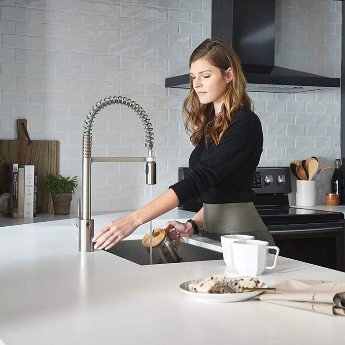 A woman pours water from a kettle into a sink in a modern kitchen, featuring white tiles, wooden accents, and kitchen utensils nearby.