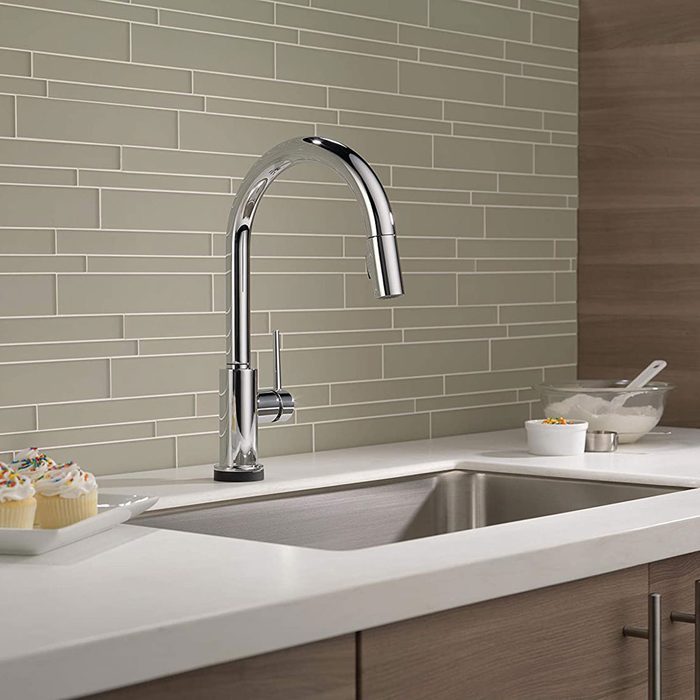 A sleek, chrome kitchen faucet extends over a stainless steel sink; beige tile backdrop complements a countertop with cupcakes and bowls of ingredients nearby.