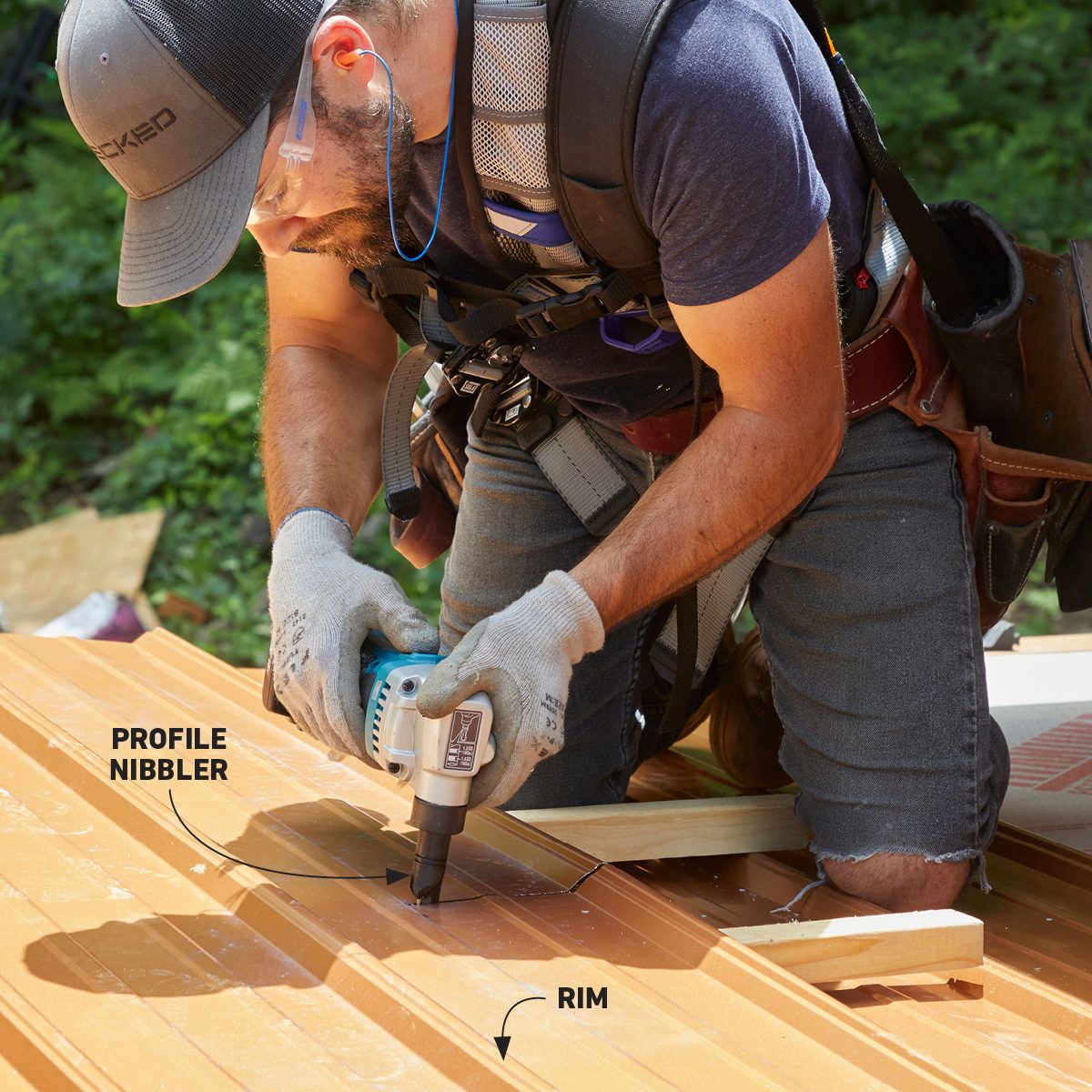 A worker uses a profile nibbler tool to cut a metal sheet while kneeling on orange roofing material in a green outdoor setting.