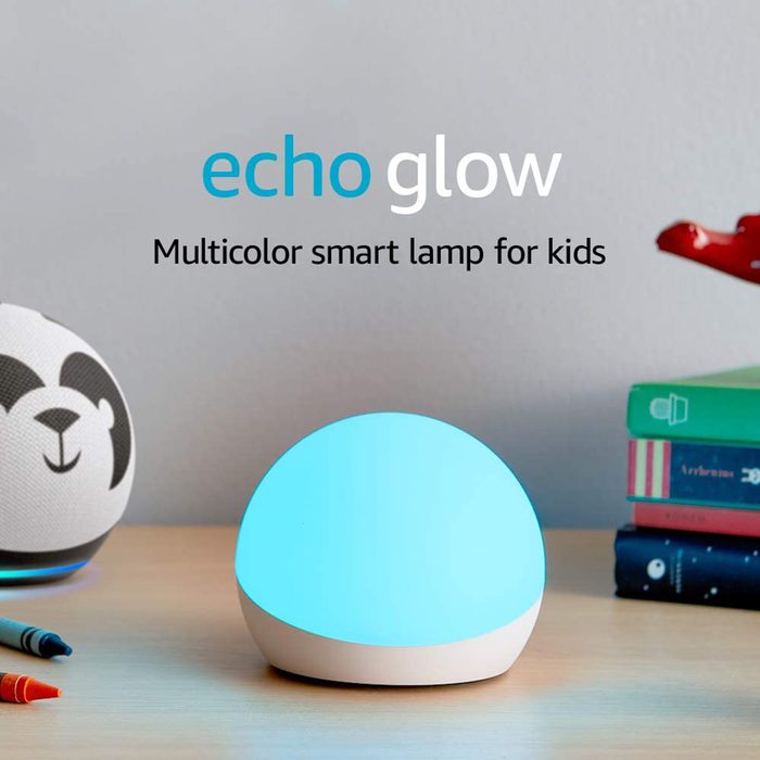 A round, blue smart lamp glows on a table, surrounded by colorful crayons, a panda speaker, and stacked books against a light-colored wall.