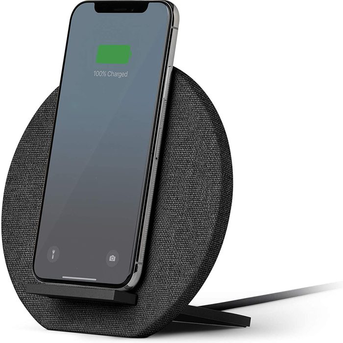 A smartphone is charging on a circular, fabric-covered wireless charger, displaying a full battery indicator in a minimalist, modern setting.