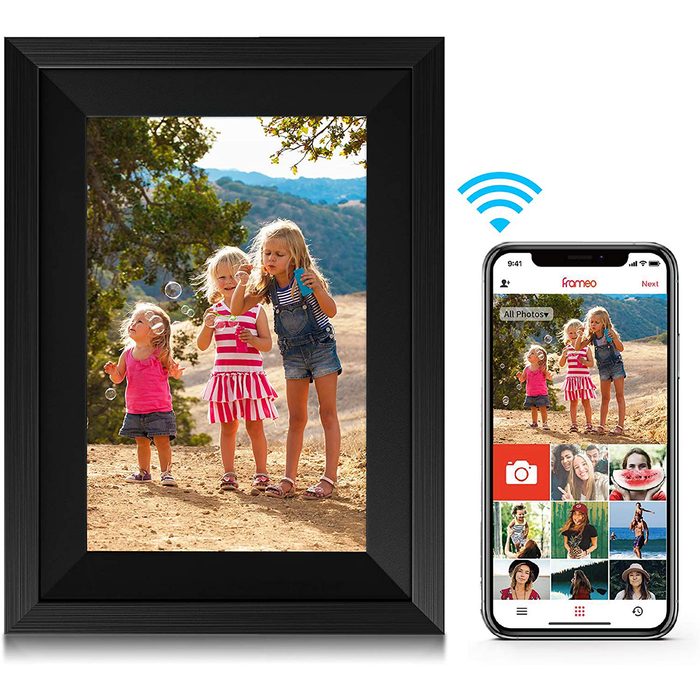 Three children blow bubbles while standing outdoors, surrounded by trees and hills, with a digital photo frame displaying their image and a smartphone nearby.