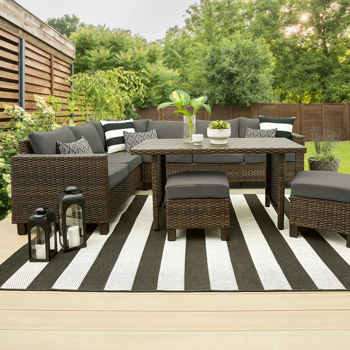 A wicker seating area with gray cushions surrounds a table on a striped rug, set in a lush outdoor space with greenery and wooden fencing.