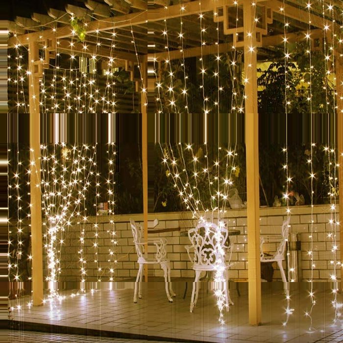 String lights hang gracefully from a wooden pergola, illuminating a patio with white chairs and a tiled floor, creating a warm, inviting atmosphere at night.