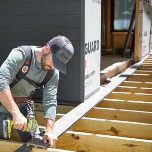 A carpenter uses a power drill to secure a board onto a wooden deck frame, surrounded by construction materials and a partially built structure.