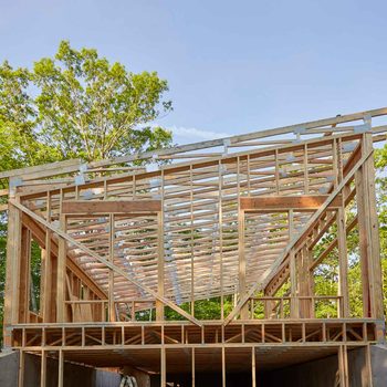 A wooden structure is being built, showcasing framed walls and rafters under a clear blue sky, surrounded by green trees.
