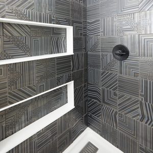 A shower wall features geometric patterned tiles in shades of black and gray, while white shelves provide storage in the modern bathroom setting.