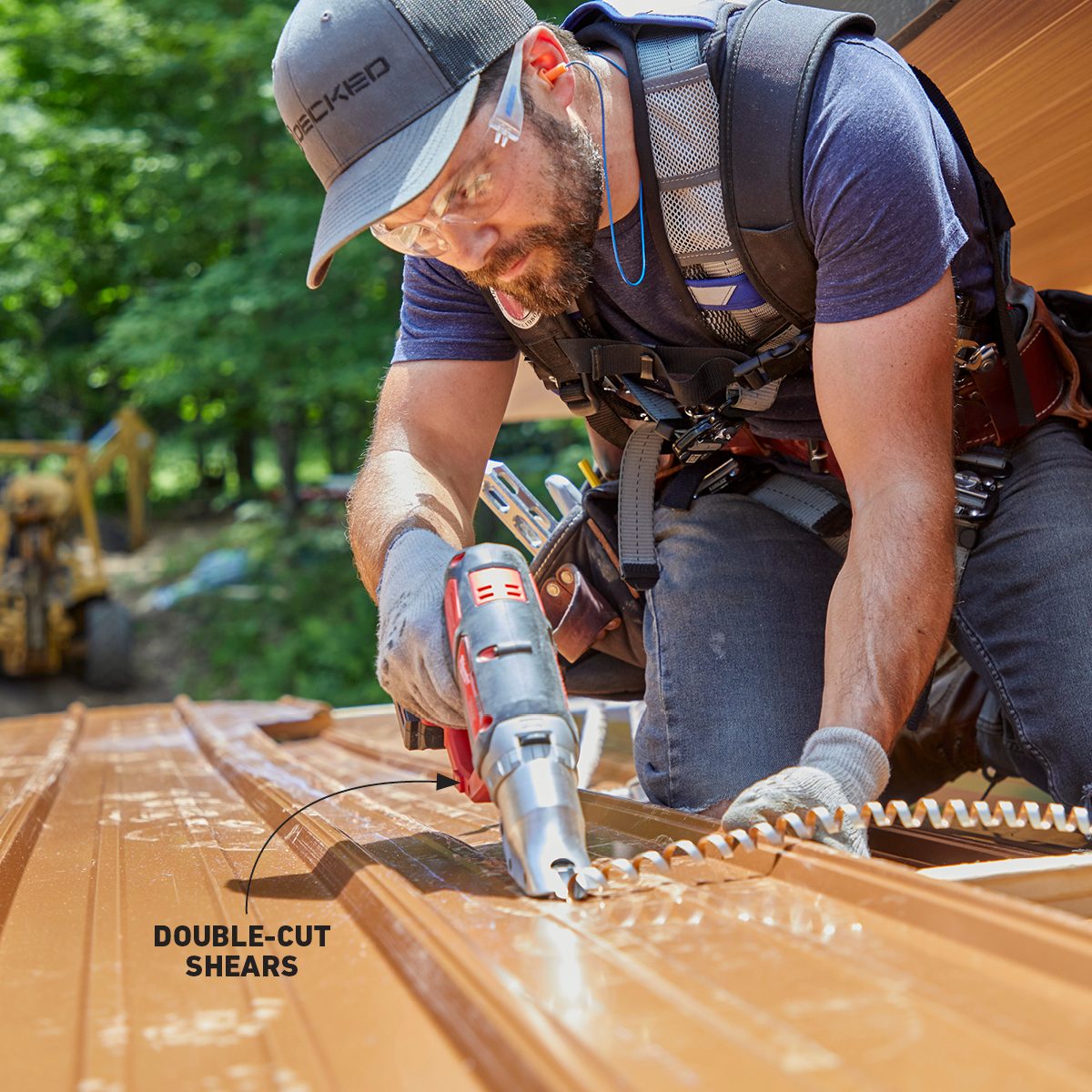 A man uses double-cut shears to cut metal roofing sheets while kneeling on a sunny construction site surrounded by trees and equipment.