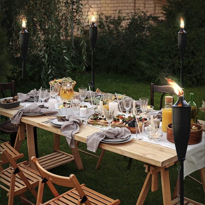 A long wooden table is set outdoors, adorned with food, drinks, and lit torches, creating an inviting atmosphere for a gathering in a garden.