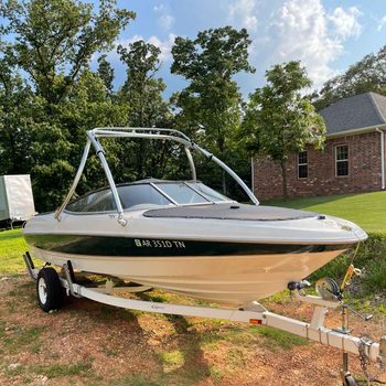 A boat with a sleek design is parked on a gravel area, surrounded by trees and grass, near a brick house under a partly cloudy sky.