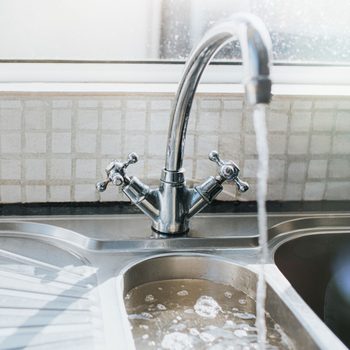 A shiny faucet releases water into a stainless steel sink, creating bubbles, against a backdrop of a window and tiled wall.
