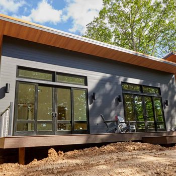 A modern house with large windows features a wooden deck and patio furniture, surrounded by trees under a partly cloudy sky.