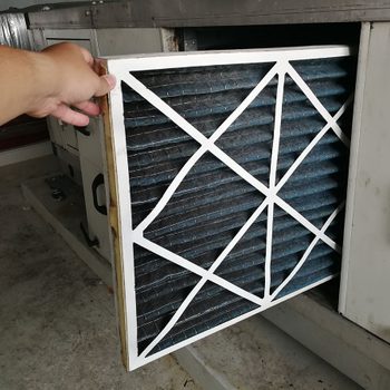 A hand holds a rectangular air filter, removing it from a machine in a clean, industrial setting with concrete flooring and metal equipment nearby.