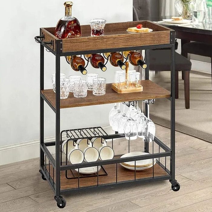 A wooden bar cart holds bottles, glasses, and plates, providing a mobile setup for serving drinks in a modern dining area with a neutral design.