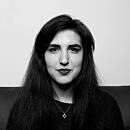 A woman with long dark hair sits facing the camera, displaying a neutral expression, against a simple, brightly lit background.