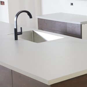 A black faucet is positioned over a square sink on a light-colored countertop, adjacent to a wooden surface, in an unfinished kitchen space.