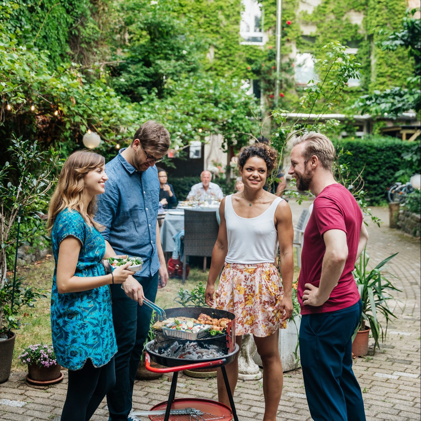 A group of four people engages around a barbecue grill in a green garden, chatting and preparing food while others dine at a nearby table.