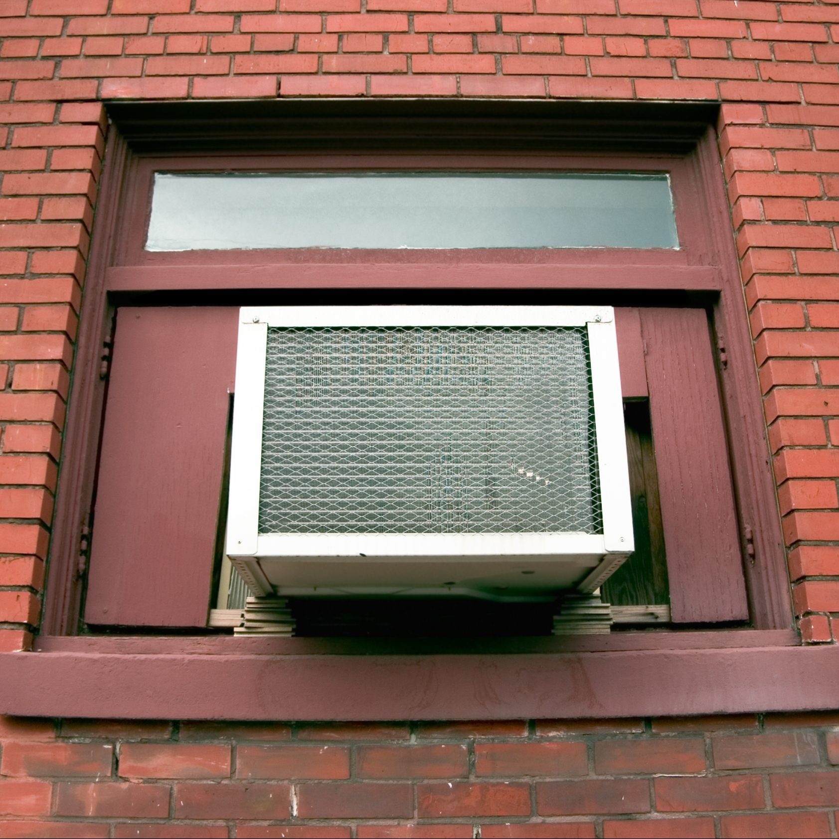 An air conditioning unit protrudes from a window, drawing in airflow, set against a brick wall with a maroon frame and a small upper window.