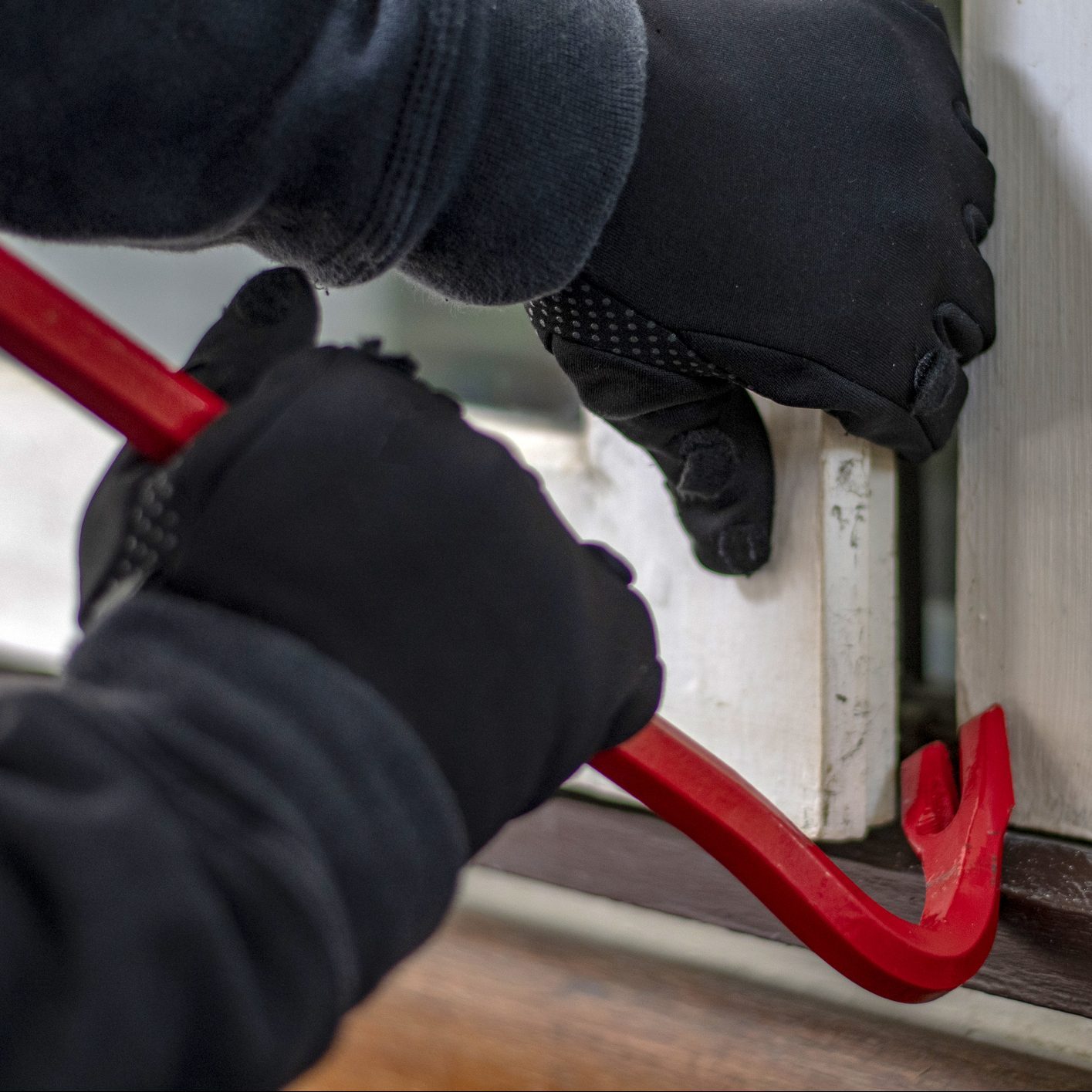 Gloved hands use a red crowbar to pry open a window in a residential setting, suggesting an attempt to gain unauthorized entry.
