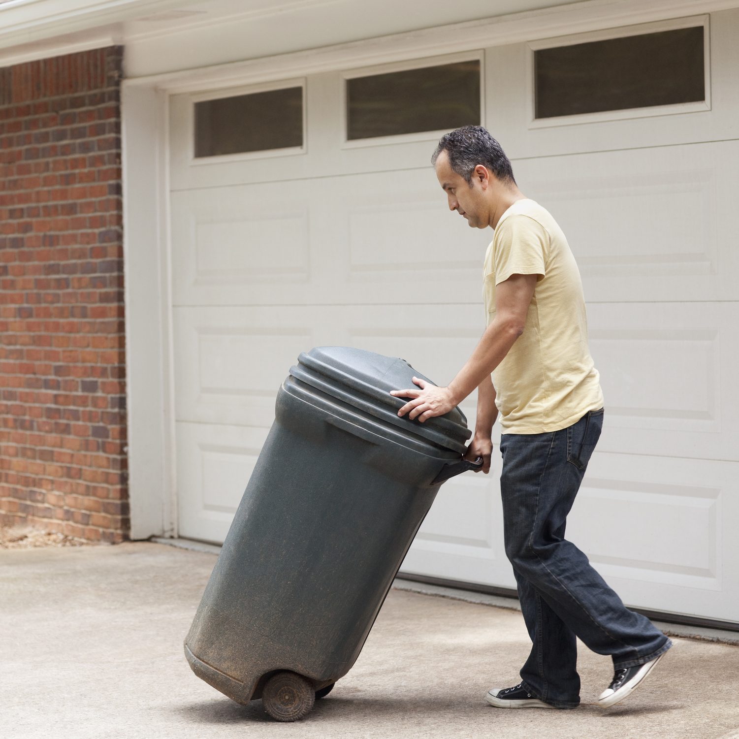 A man pushes a large, dirty trash can towards a garage, with a brick wall and a closed garage door in the background.