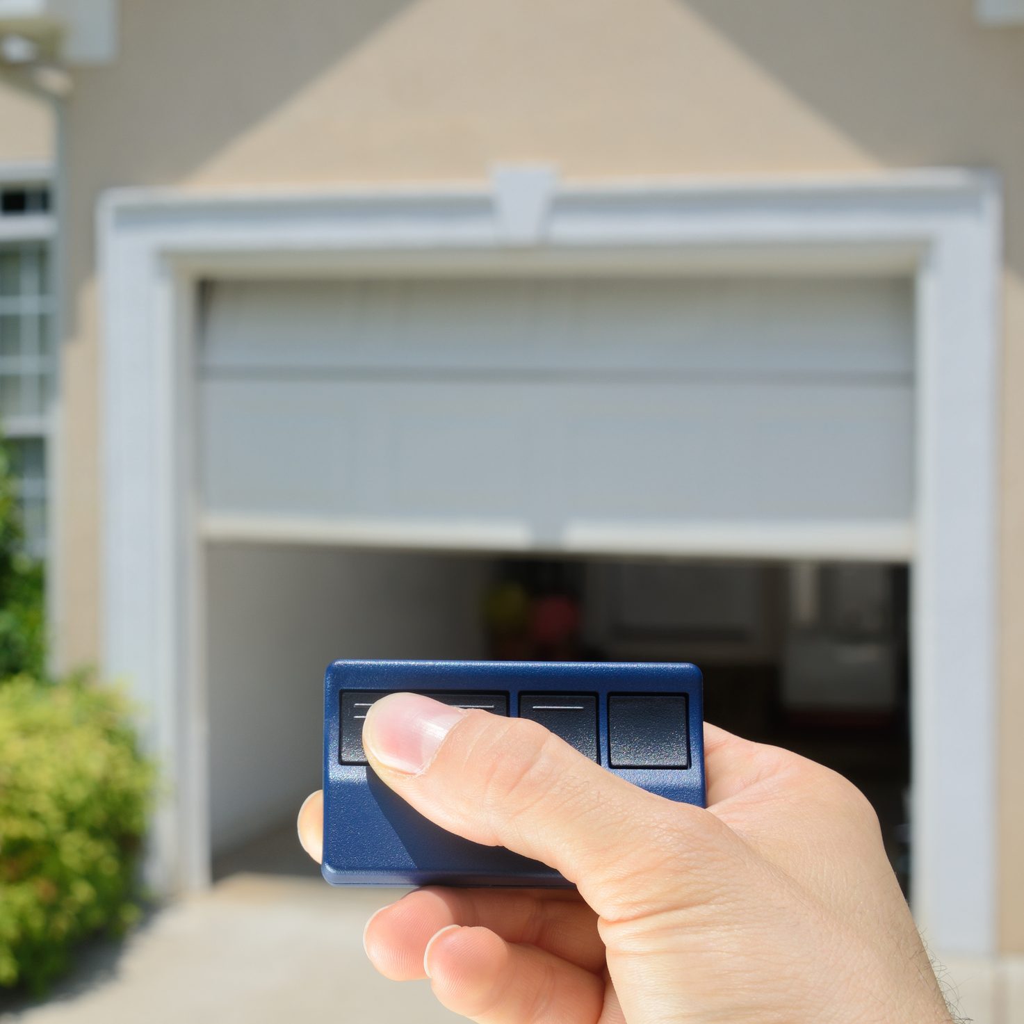 A hand holds a blue garage door remote, pressing a button to open the garage door of a light-colored house surrounded by greenery.
