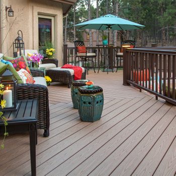 A cozy deck features wicker furniture, colorful cushions, and decorative tables. Flowers and candles add ambiance, while a turquoise umbrella provides shade in a wooded setting.