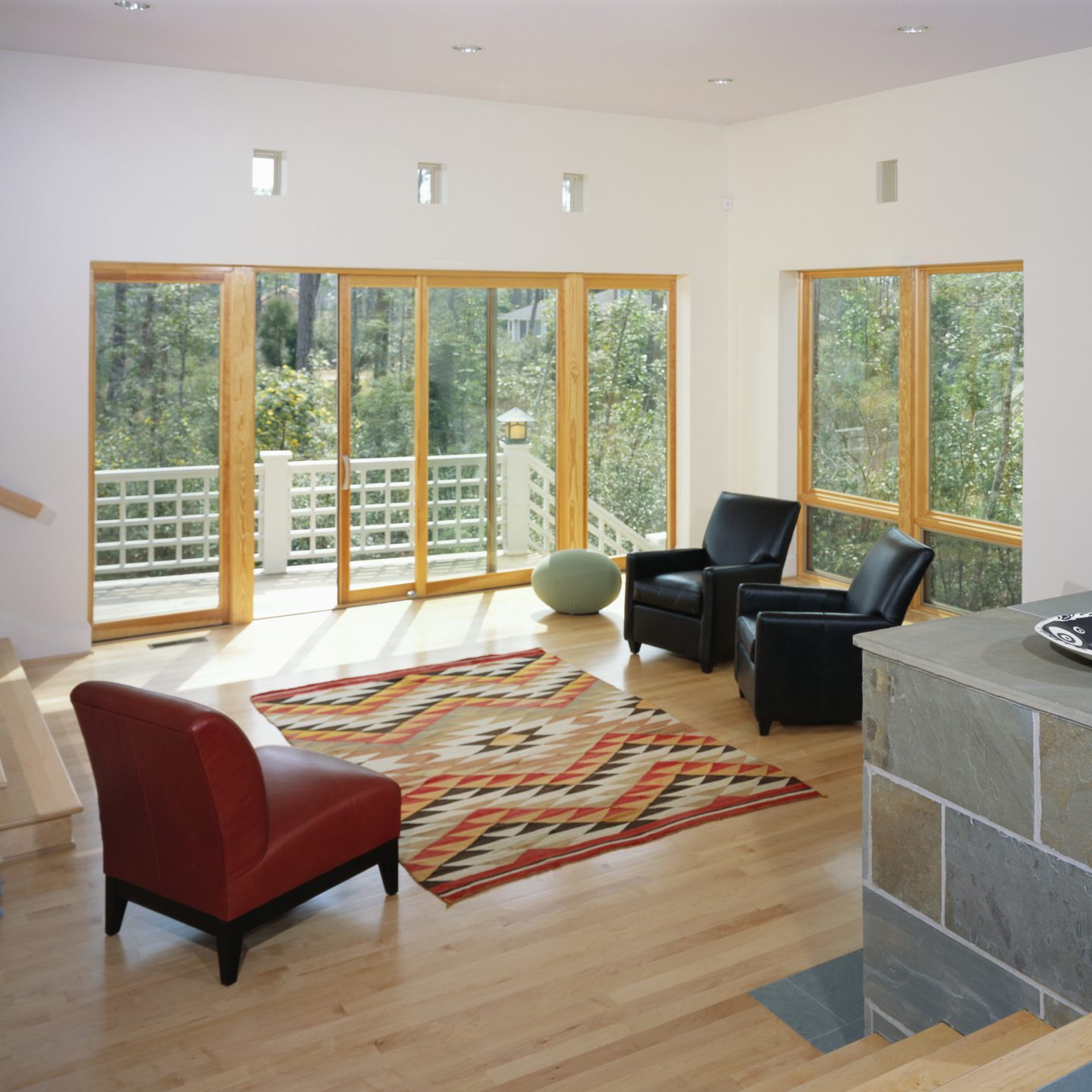 A red chair and two black chairs sit on a patterned rug in a sunny living room, with large windows overlooking greenery.