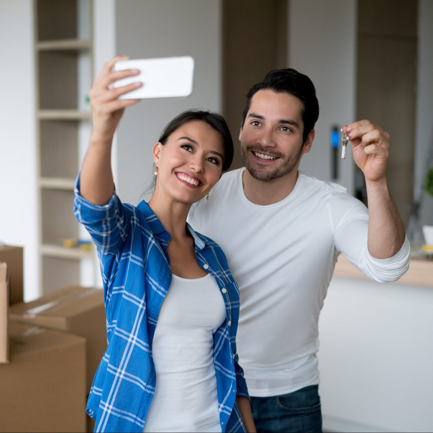 Two smiling young adults take a selfie while holding keys. They stand in a room with unpacked boxes, indicating a recent move.