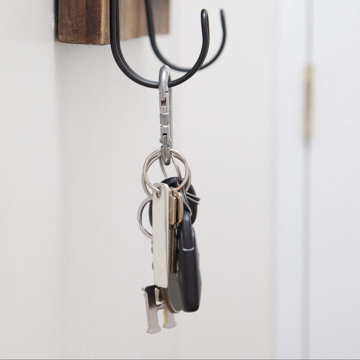 A metal keyring hangs from a black hook on a wooden wall, displaying multiple keys, including a car key, against a plain white background.