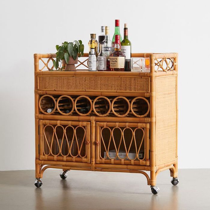 A rattan bar cart displays several bottles and a potted plant on top, while featuring wine storage and two cabinets below, set against a neutral wall.