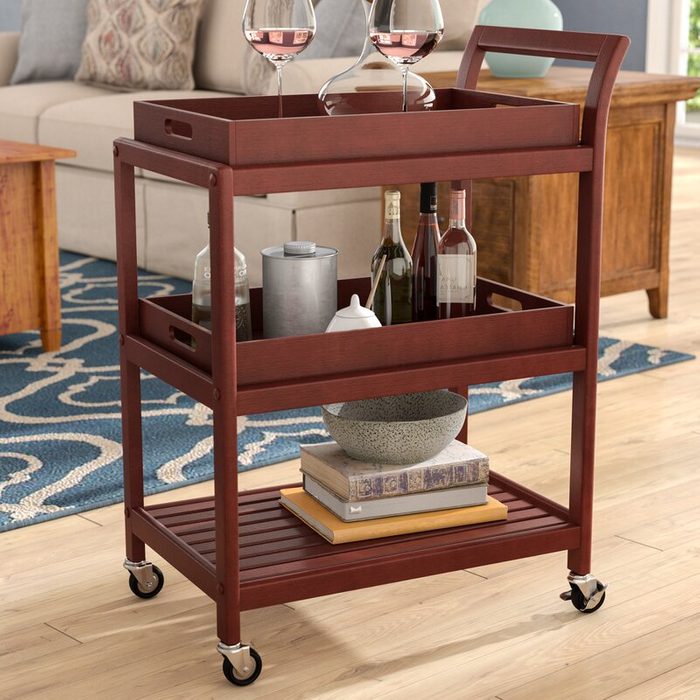 A wooden serving cart holds wine glasses and bottles, a decanter, and a bowl, positioned on a patterned rug in a cozy living room setting.