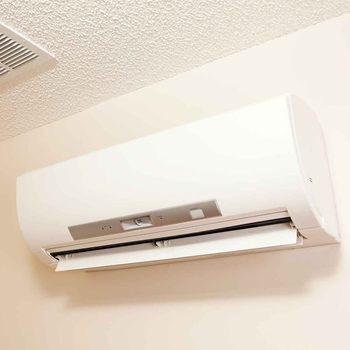 A white air conditioning unit is mounted on a wall, blowing cool air into a neutral-colored room with textured ceilings.