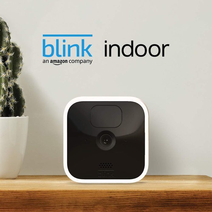 A security camera sits on a wooden shelf next to a potted cactus, against a light-colored wall, labeled with 