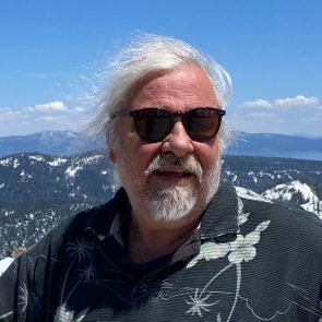 A man with long, white hair and sunglasses smiles against a backdrop of mountains and blue sky, featuring snow-capped peaks and distant lakes.
