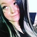 A person with black and silver hair is looking slightly to the side, displaying a playful expression in a casual indoor setting.