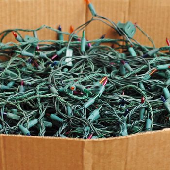 A tangled mass of green Christmas lights rests inside a cardboard box, with multicolored bulbs peeking through the wires, suggesting disorganization.