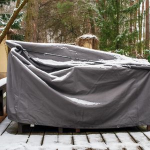 A gray cover obscures an outdoor furniture set, resting on a snow-dusted wooden deck surrounded by trees and a partially visible garden.