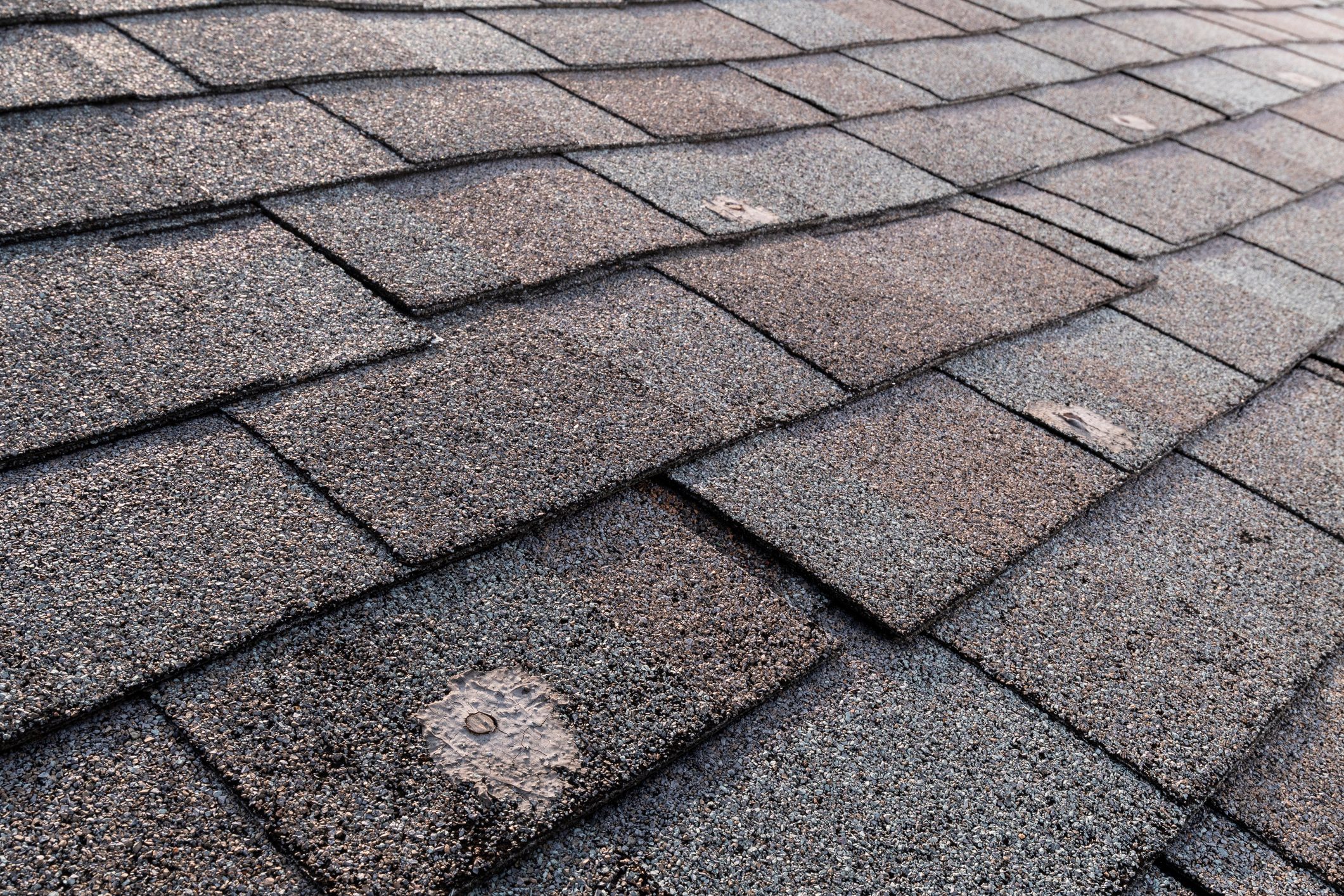 Shingles cover a sloped rooftop, overlapping in a pattern, reflecting light, with a textured surface indicating wear and weather exposure.
