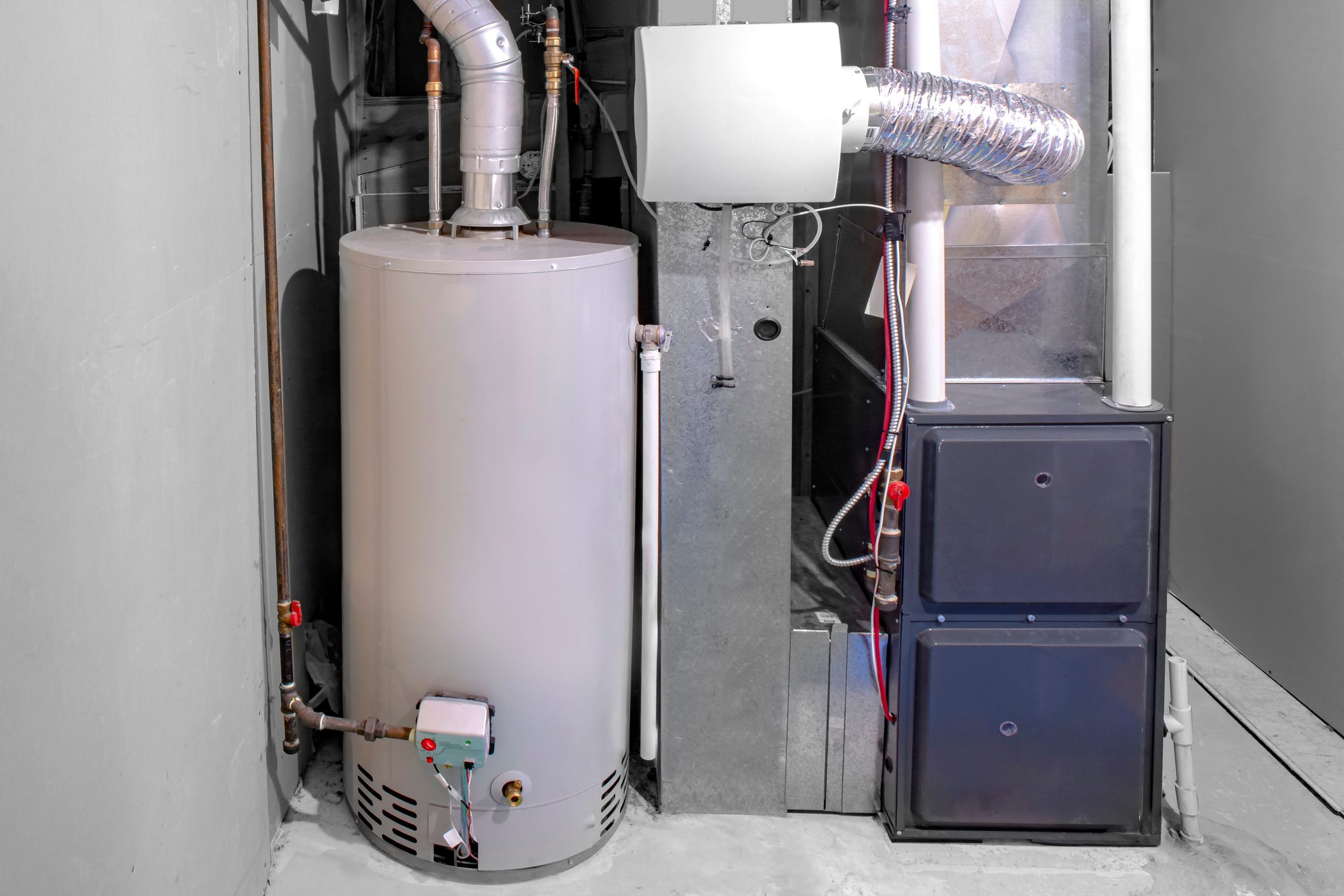 A water heater stands next to a vented unit in a basement, connected by pipes and positioned against gray walls on a concrete floor.
