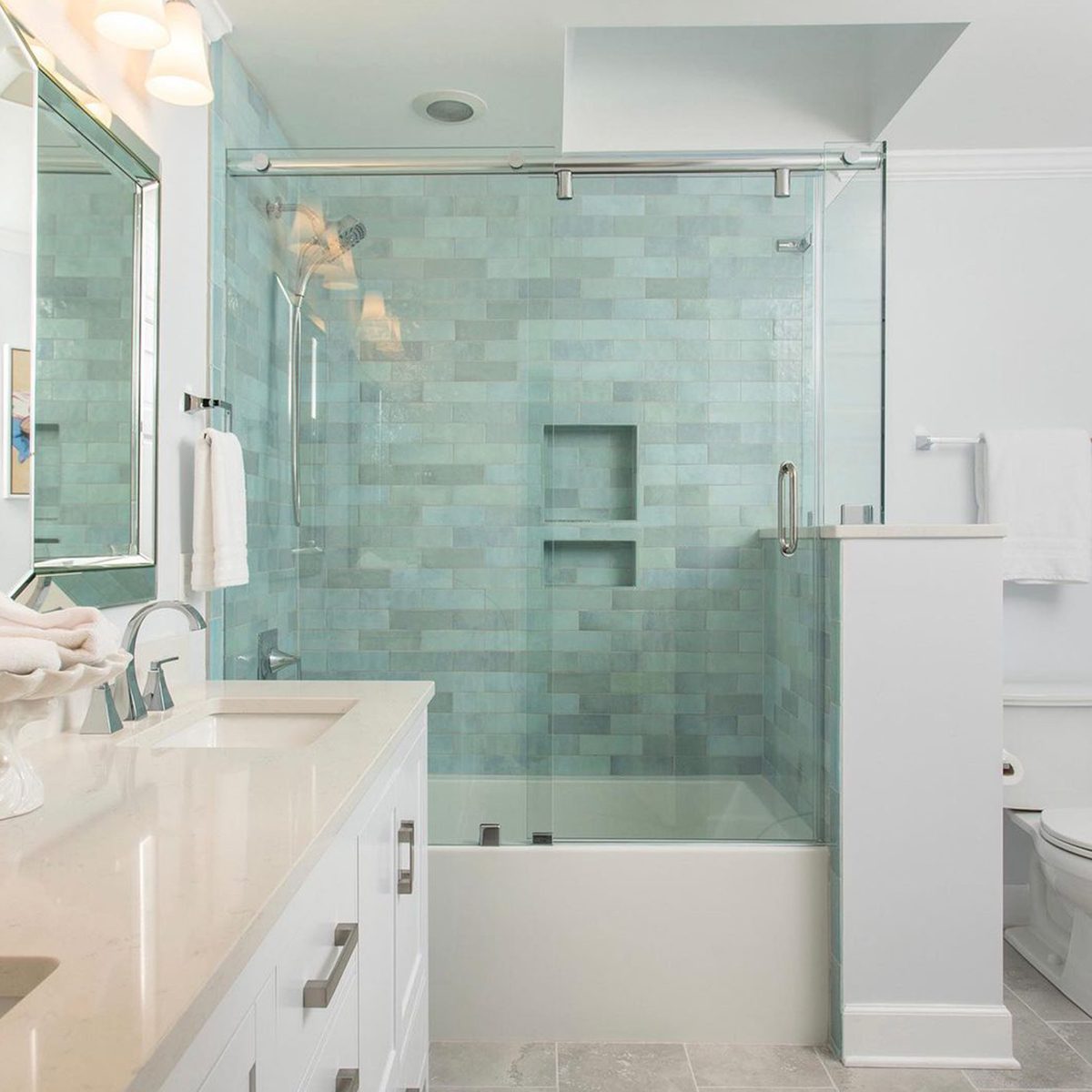 A modern bathroom features a glass shower with a light blue tiled wall, a dual-sink vanity, and white towels hanging nearby, enhancing the serene atmosphere.