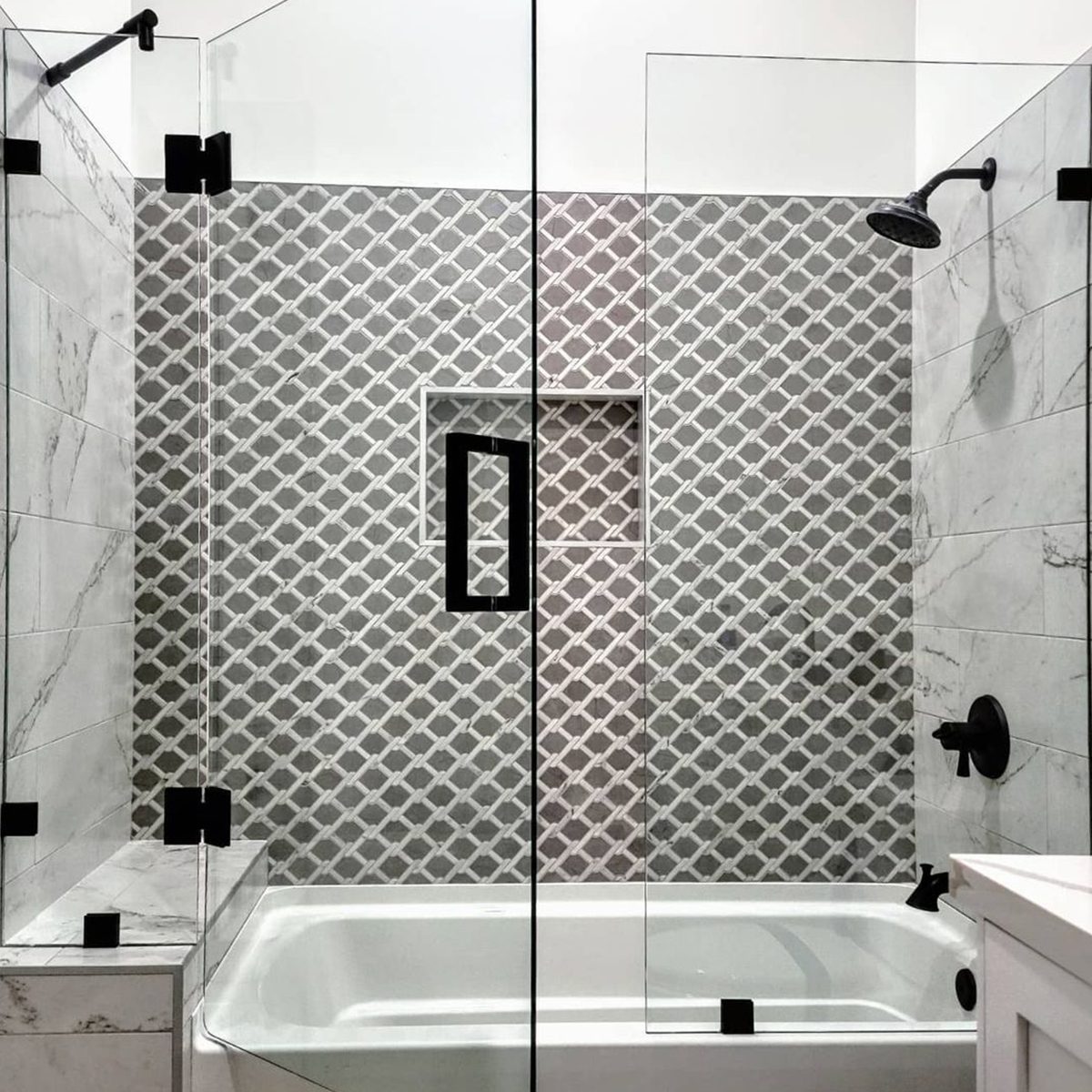 A glass shower enclosure reveals a patterned tiled wall and a bathtub below. A showerhead and fixtures complete the modern bathroom setting.