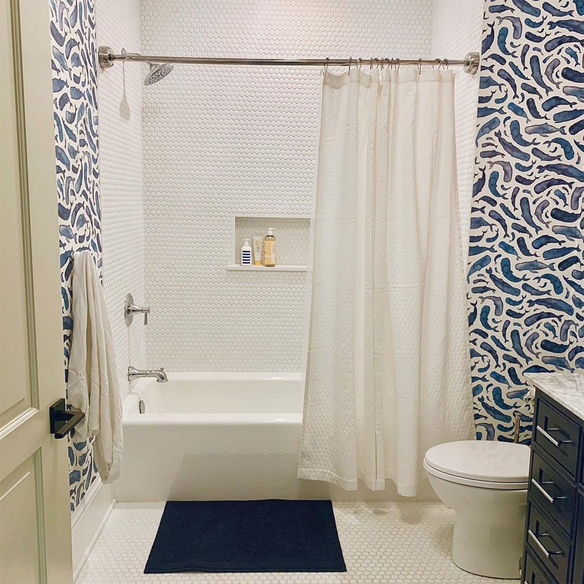 A white bathtub with a shower curtain stands in a bathroom featuring patterned blue wallpaper and a small built-in shelf with toiletries.