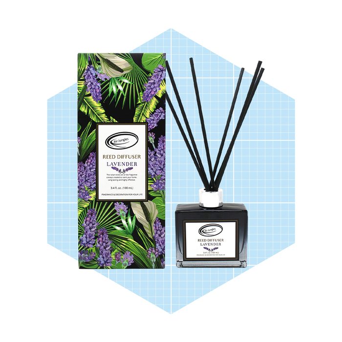 A lavender reed diffuser sits beside its decorative packaging, surrounded by a light blue geometric background. The diffuser emits fragrance for ambient enjoyment.